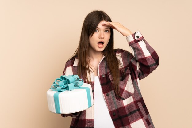 Ragazza che tiene una grande torta isolata sul beige facendo il gesto di sorpresa mentre guarda al lato