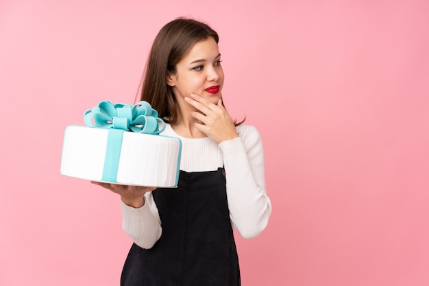 Ragazza che tiene una grande torta sul rosa che pensa un'idea e che guarda lato