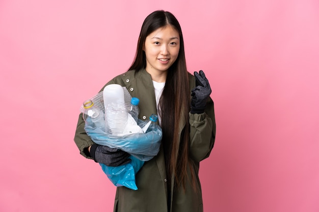 Ragazza cinese che tiene un sacchetto pieno di bottiglie di plastica da riciclare sopra fondo rosa isolato che fa gesto dei soldi