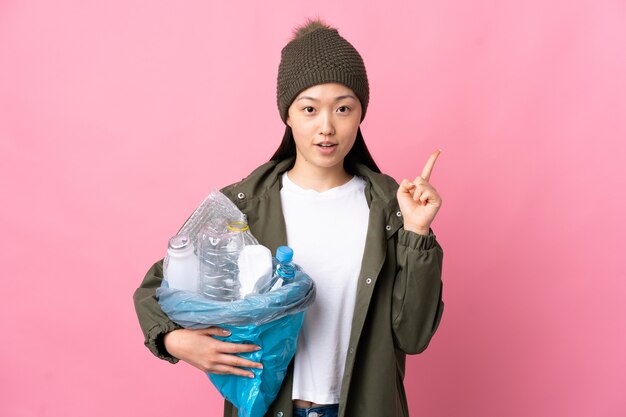 Ragazza cinese che tiene una borsa piena di bottiglie di plastica da riciclare sul rosa isolato pensando a un'idea che punta il dito verso l'alto