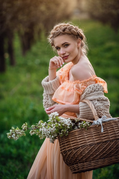 Ragazza con capelli biondi in un vestito leggero nel giardino fiorito. ragazza in un bel vestito e maglione lavorato a maglia si gode il tramonto in un giardino fiorito di pere, con un cesto di fiori