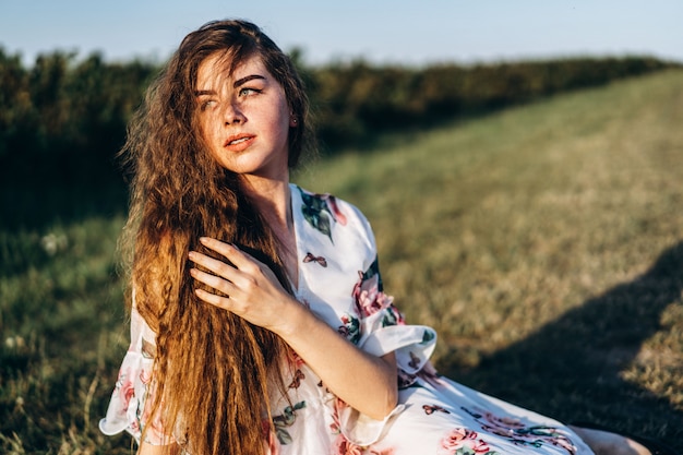 Ragazza con capelli ricci lunghi, castana, fronte in lentiggini, con trucco e occhi verdi, in vestito bianco leggero che posa per la macchina fotografica, fondo vago, natura.