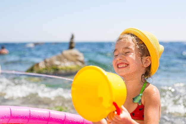 Ragazza con cappello di paglia giallo gioca con l'acqua del vento e un erogatore d'acqua in una piscina gonfiabile sulla spiaggia Prodotti indelebili per proteggere la pelle dei bambini dal sole resort al mare