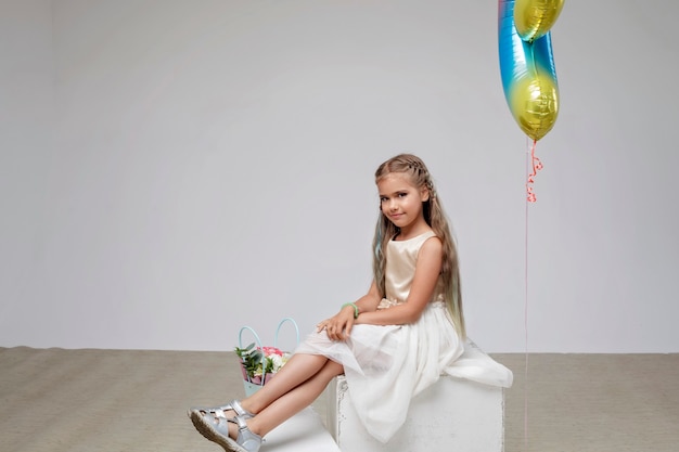 Ragazza con i capelli lunghi in abito festivo seduto vicino a bouquet di fiori sfondo bianco studio