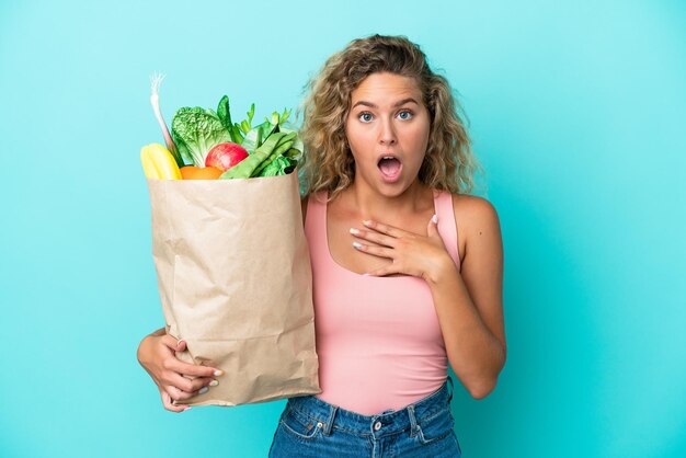 Ragazza con i capelli ricci che tiene una borsa della spesa isolata su sfondo verde sorpresa e scioccata mentre guarda a destra