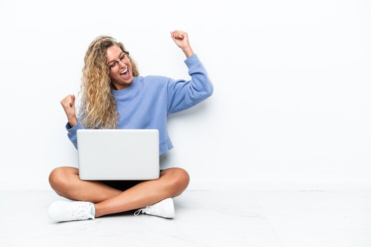 Foto ragazza con i capelli ricci con un computer portatile seduto sul pavimento che celebra una vittoria