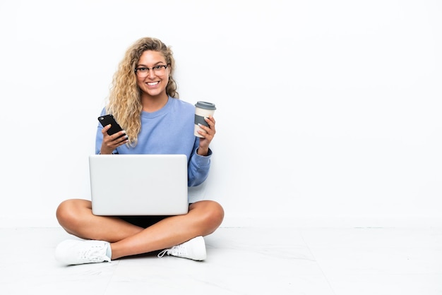 Ragazza con i capelli ricci con un computer portatile seduto sul pavimento con in mano un caffè da portare via e un cellulare