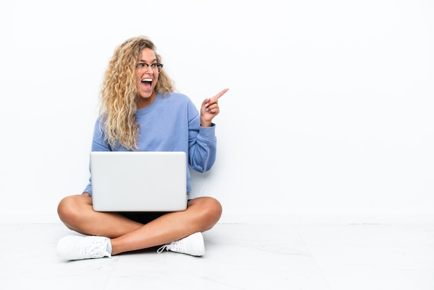 Ragazza con i capelli ricci con un laptop seduto sul pavimento che punta il dito di lato e presenta un prodotto