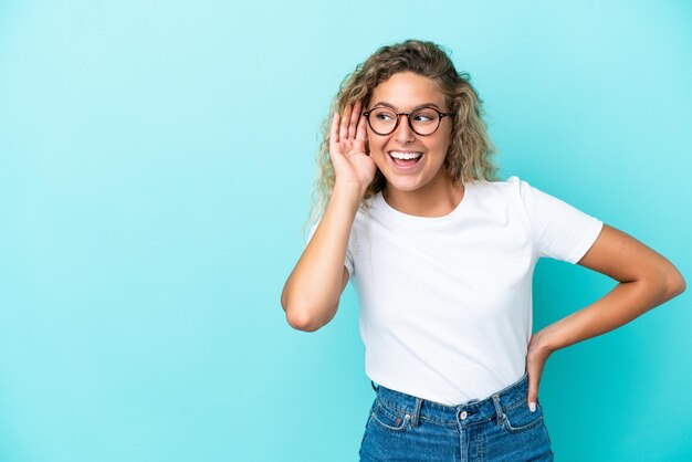 Ragazza con i capelli ricci isolata su sfondo blu che ascolta qualcosa mettendo la mano sull'orecchio