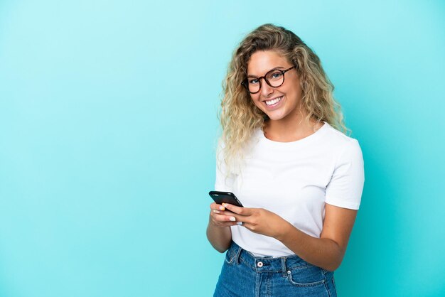 Ragazza con i capelli ricci isolata su sfondo blu che invia un messaggio con il cellulare