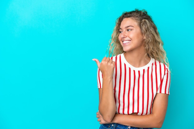 Ragazza con i capelli ricci isolata su sfondo blu che punta al lato per presentare un prodotto