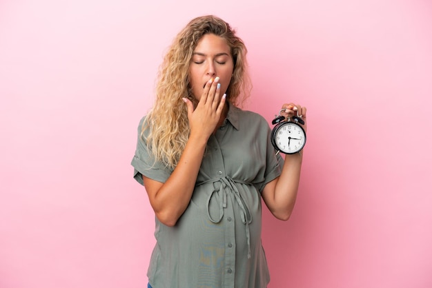 Ragazza con i capelli ricci isolata su sfondo rosa incinta e tenendo l'orologio mentre sbadiglia