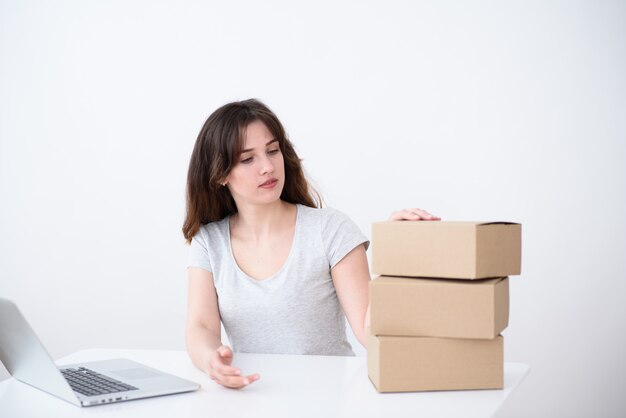 Ragazza con i capelli, una maglietta grigia seduto a un computer portatile e guardando una pila di scatole di cartone. Servizio di consegna online