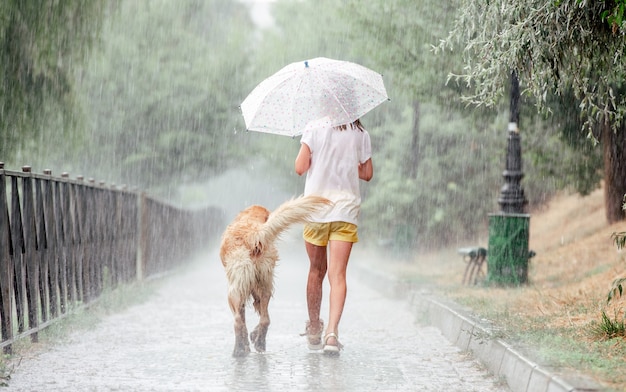 Ragazza con il cane golden retriever durante la pioggia che cammina bagnata sotto l'ombrello fuori. Preteen kid con doggy pet in giorno di pioggia indietro ritratto