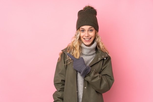 Ragazza con il cappello di inverno isolato sulla parete rosa che indica dito il lato