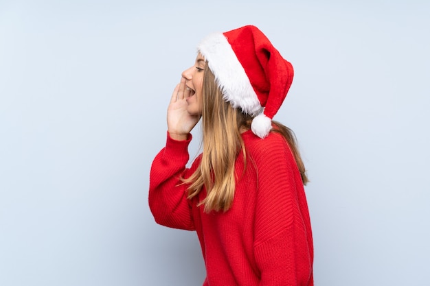 Ragazza con il cappello di natale sopra la parete blu isolata che grida con la bocca spalancata