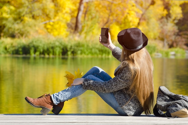 Ragazza con il cappello seduto sul molo e fa selfie. Giornata di sole autunnale. Vista laterale