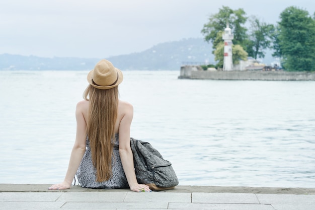 Ragazza con il cappello seduto sul molo. Mare e piccolo faro in lontananza. Vista posteriore