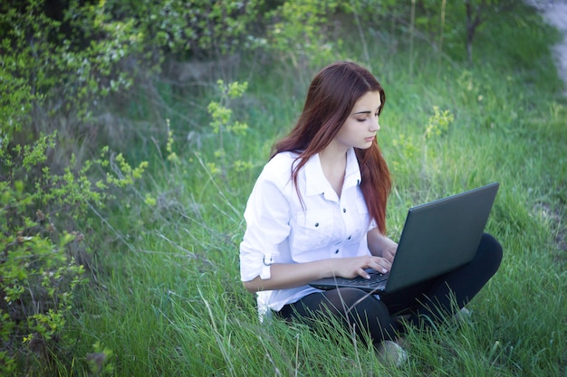Ragazza con il computer portatile in natura, ragazza che si siede sull&#39;erba e che utilizza computer portatile che funziona in natura