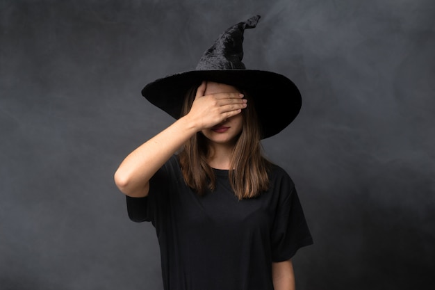 Ragazza con il costume da strega per feste di halloween sopra isolato muro scuro che copre gli occhi a mano