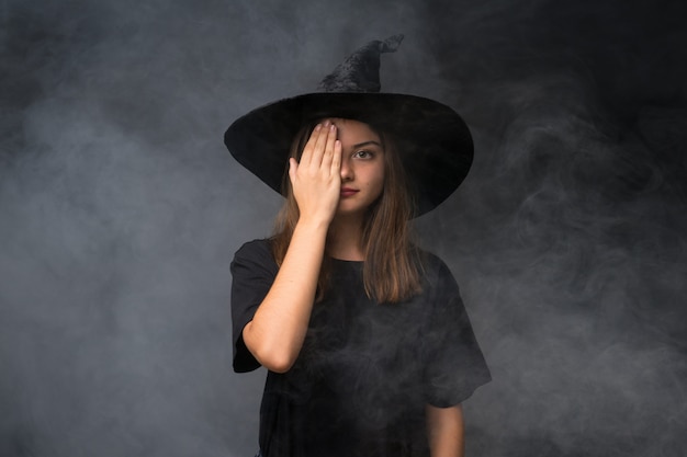 Ragazza con il costume della strega per le feste di Halloween sopra la parete scura isolata che copre a mano un occhio