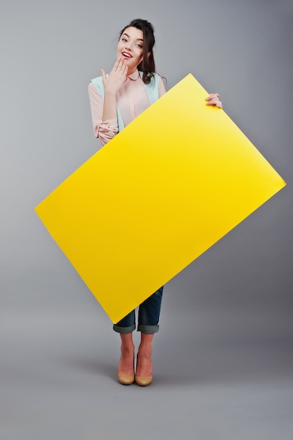 Ragazza con il fronte sorpreso tiene la carta in bianco gialla. Carta in bianco sorridente di giovane manifestazione sorridente della donna. Ragazza con il ritratto dei capelli lunghi isolato su fondo grigio.