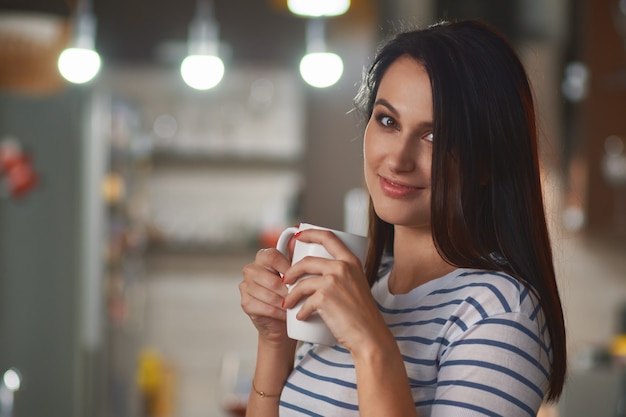 Ragazza con la tazza bianca nelle mani sulla cucina sfondo sfocato