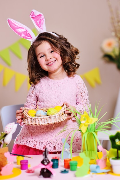 Ragazza con le orecchie di coniglio che tengono un cestino di Pasqua con le uova colorate verniciate e che sorridono sulla superficie della decorazione di Pasqua