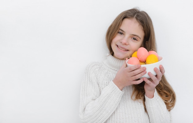 ragazza con le uova di Pasqua rosa e gialle nelle sue mani, che indossa un maglione bianco contro un muro bianco