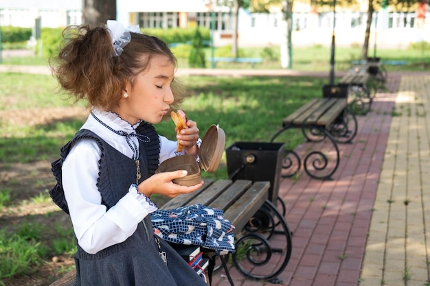 Ragazza con lo zaino che mangia un panino confezionato in una scatola di panini vicino alla scuola Uno spuntino veloce con un panino con cibo malsano pranzo da scuola Ritorno a scuola Istruzione classi della scuola primaria 1 settembre