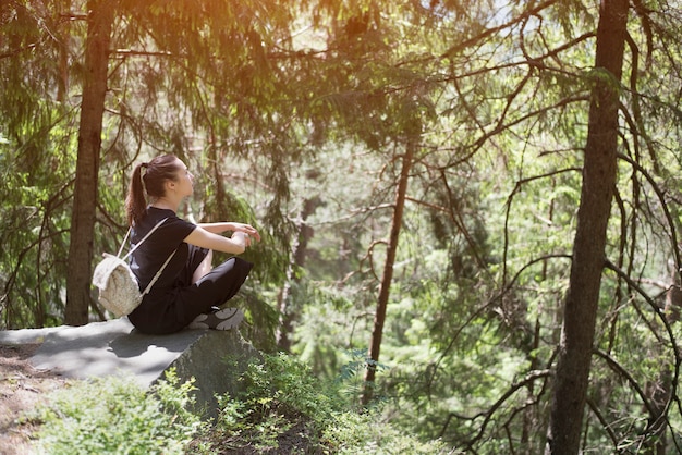 Ragazza con lo zaino che si siede su una roccia nella foresta. Giornata di sole estivo