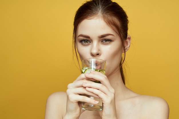 Ragazza con un bicchiere d'acqua con un cetriolo su uno studio di sfondo giallo