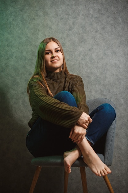 Ragazza con un maglione verde e jeans su una sedia grigia e sorridente