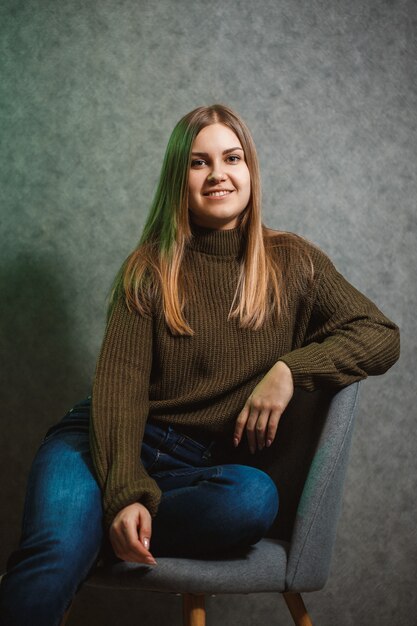 Ragazza con un maglione verde e jeans su una sedia grigia e sorridente