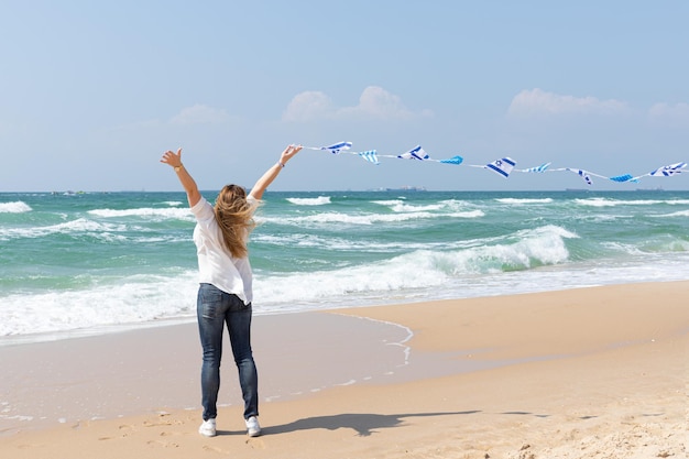 Ragazza con una ghirlanda di bandiere di Israele mostra un dito un segno eccellente in riva al mare Vacanza patriottica Giorno dell'Indipendenza Israele Yom Ha'atzmaut concept