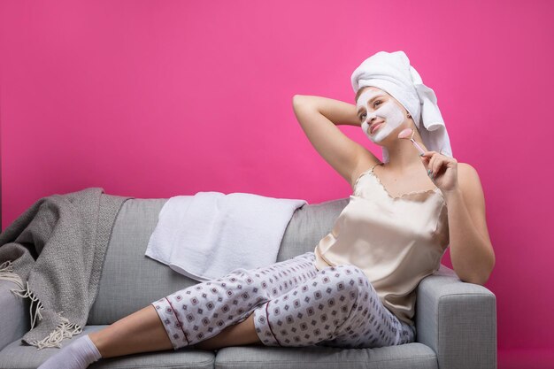 Ragazza con una maschera cosmetica sul viso in un asciugamano bianco. Si siede sul divano.