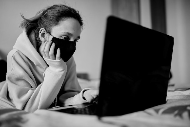 Ragazza con una maschera nera lavora su un laptop a casa in isolamento foto in bianco e nero