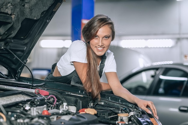 Ragazza con una tuta nera e una maglietta bianca vicino al cappuccio aperto. La ragazza nel garage sta sorridendo alla macchina fotografica