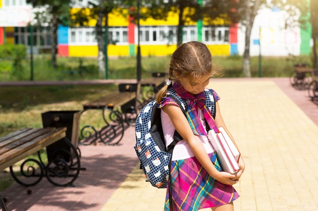 Ragazza con uno zaino e una pila di libri vicino alla scuola. Torna a scuola, il bambino è stanco, libri di testo pesanti. Istruzione, classi della scuola primaria, inizio anno scolastico, 1 settembre