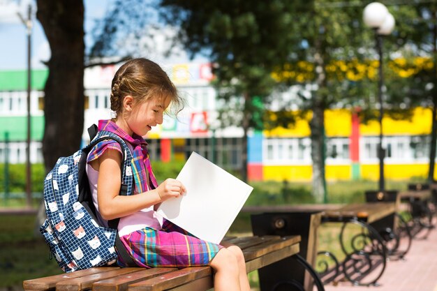 Ragazza con uno zaino seduto su una panchina e leggendo un libro vicino alla scuola. Ritorno a scuola, orario delle lezioni, diario con i voti. Istruzione, classi della scuola primaria, 1 settembre