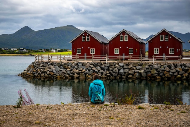 ragazza con uno zaino si siede in riva al mare con vista sui cottage rossi e sulle montagne, le isole lofoten