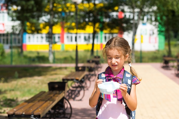 Ragazza con uno zaino vicino alla scuola dopo le lezioni si toglie una maschera medica, infelice