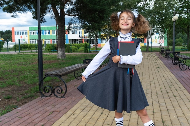 Ragazza con zaino uniforme scolastica con fiocchi bianchi e pila di libri vicino alla scuola Ritorno a scuola felice allievo libri di testo pesanti Istruzione classi della scuola primaria 1 settembre