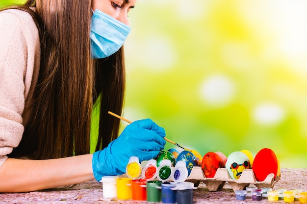 Ragazza concentrata caucasica in maschera medica e guanti usa e getta decora le uova fatte a mano per le vacanze pascal