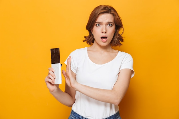 Ragazza confusa che indossa una t-shirt in piedi isolata sul muro giallo, con in mano una barretta di cioccolato