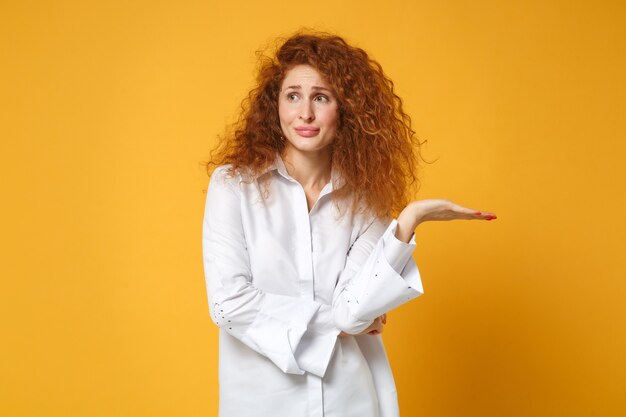 Ragazza confusa giovane donna dai capelli rossi in camicia bianca casual in posa isolata sul muro giallo arancione