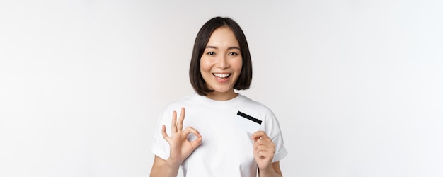 Ragazza coreana sorridente che mostra il segno giusto e che consiglia la carta di credito della banca dello spazio della copia che sta in maglietta sopra fondo bianco