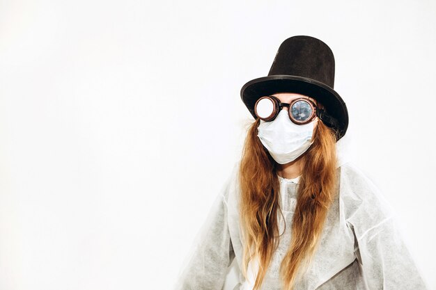Ragazza creativa in un abito bianco, maschera medica, occhiali e un cappello su un muro bianco. Autoisolamento durante la quarantena del coronavirus. Epidemic covid-2019.