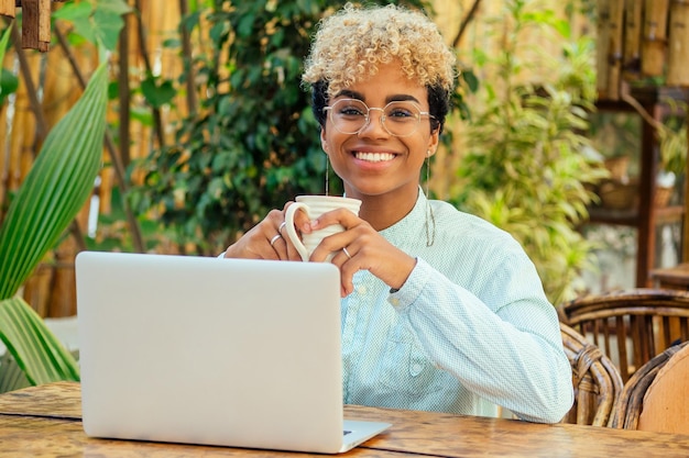 Ragazza d'affari afroamericana con capelli biondi ricci carini che usa il computer portatile e fa una pausa per una tazza di caffè al bar del giardino tropicale estivo