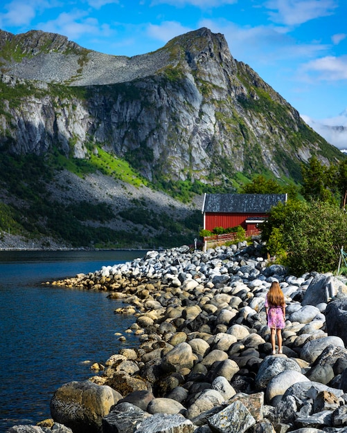 ragazza dai capelli lunghi in abito viola si erge sulle rocce con una casa rossa e imponenti montagne sullo sfondo